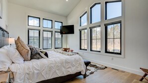 Une chambre spacieuse avec un grand lit, de nombreuses fenêtres, un téléviseur fixé au mur et un plancher en bois.