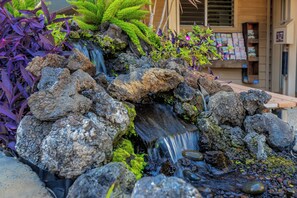 A small waterfall feature with rocks and plants, surrounded by a building with a window and a sign. - Hale Pau Hana #707 Ocean Front, Central AC, Oceanfront Complex (Kihei)