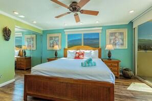 A bedroom with a wooden bed, two bedside tables, a ceiling fan, and large windows with a mountain view. - Hale Pau Hana #707 Ocean Front, Central AC, Oceanfront Complex (Kihei)