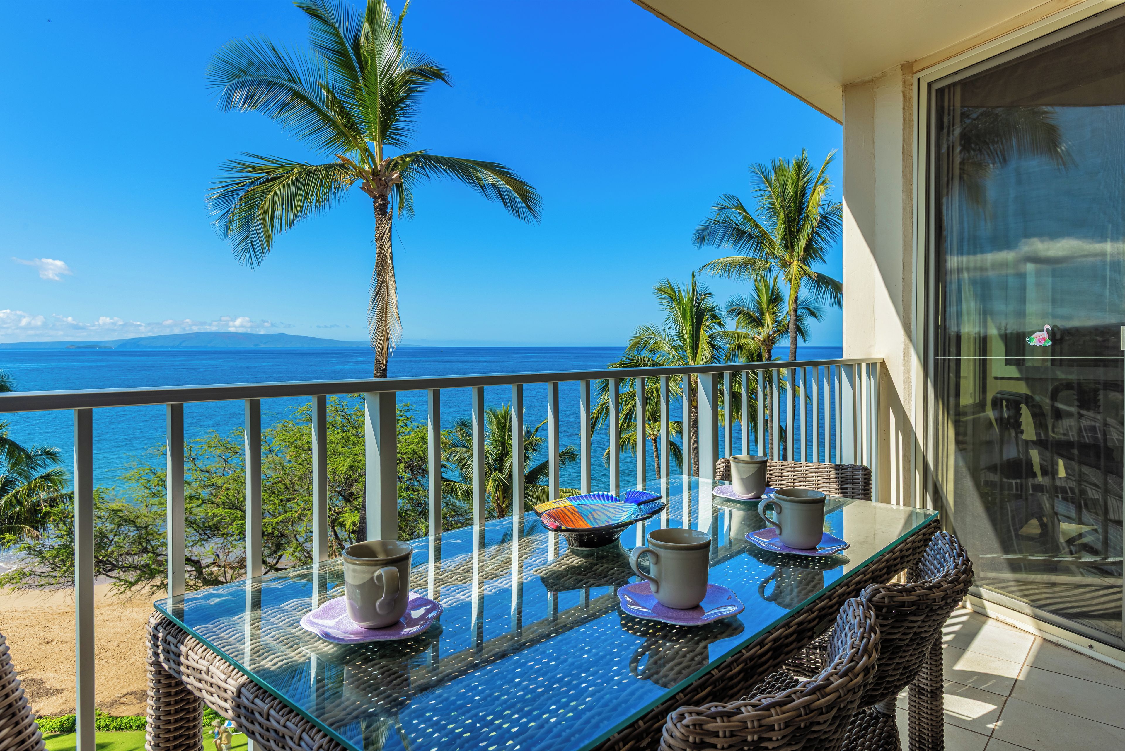 Un balcon avec une table en verre, des chaises en osier et une vue sur l’océan et les palmiers.