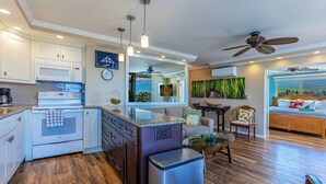Una cocina moderna con microondas, horno y refrigerador de acero inoxidable. Hay un comedor con mesa y sillas, y una sala de estar con sofá y vistas al exterior.