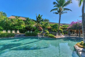 A clear pool surrounded by lush greenery and palm trees, with lounge chairs and a building in the background.