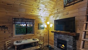 Interior acogedor de una cabaña con chimenea de piedra, un televisor montado arriba, una mesa de comedor con sillas y una ventana con vistas a árboles.