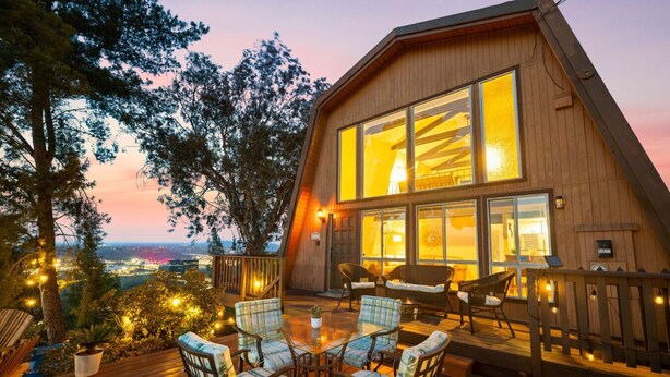Una cabaña de madera con ventanas amplias, una terraza con mobiliario exterior y vistas a la ciudad al anochecer.