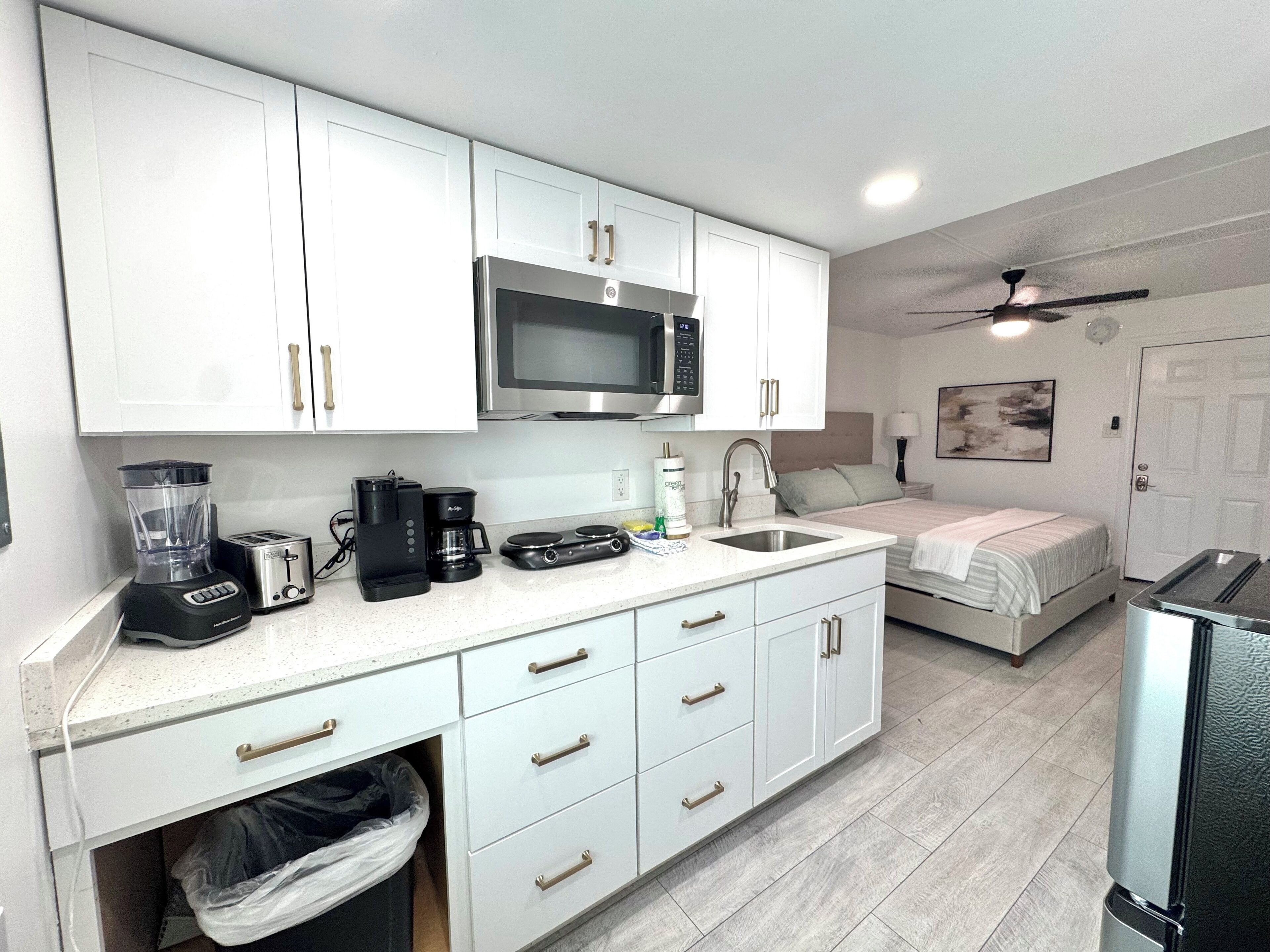 Cocina compacta con armarios blancos, microondas, cafetera y lavamanos.