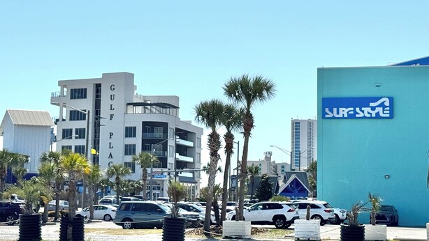Een parkeerplaats met meerdere auto's, palmbomen en gebouwen, waaronder een winkel in surfen.