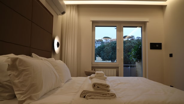 A hotel room with a bed, a window with a view of trees and buildings, and a wall-mounted air conditioning unit. - The Romex (Rome)