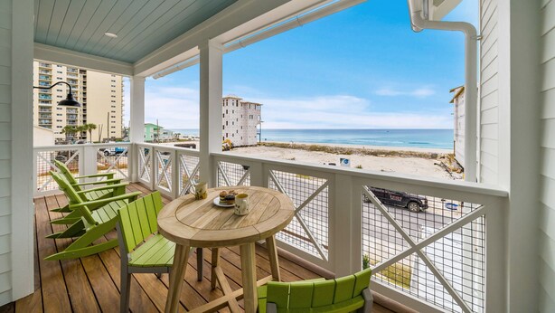 A covered outdoor area with a wooden deck, green chairs, and a table with a plate and mug. A view of the beach and ocean is visible through the railing.
