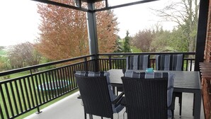 Une terrasse extérieure avec une table et des chaises, une rambarde et une vue sur des arbres et une pelouse.