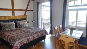 Une chambre avec un grand lit, une tête de lit en bois, une table avec des chaises et une vue sur l’extérieur par une fenêtre.