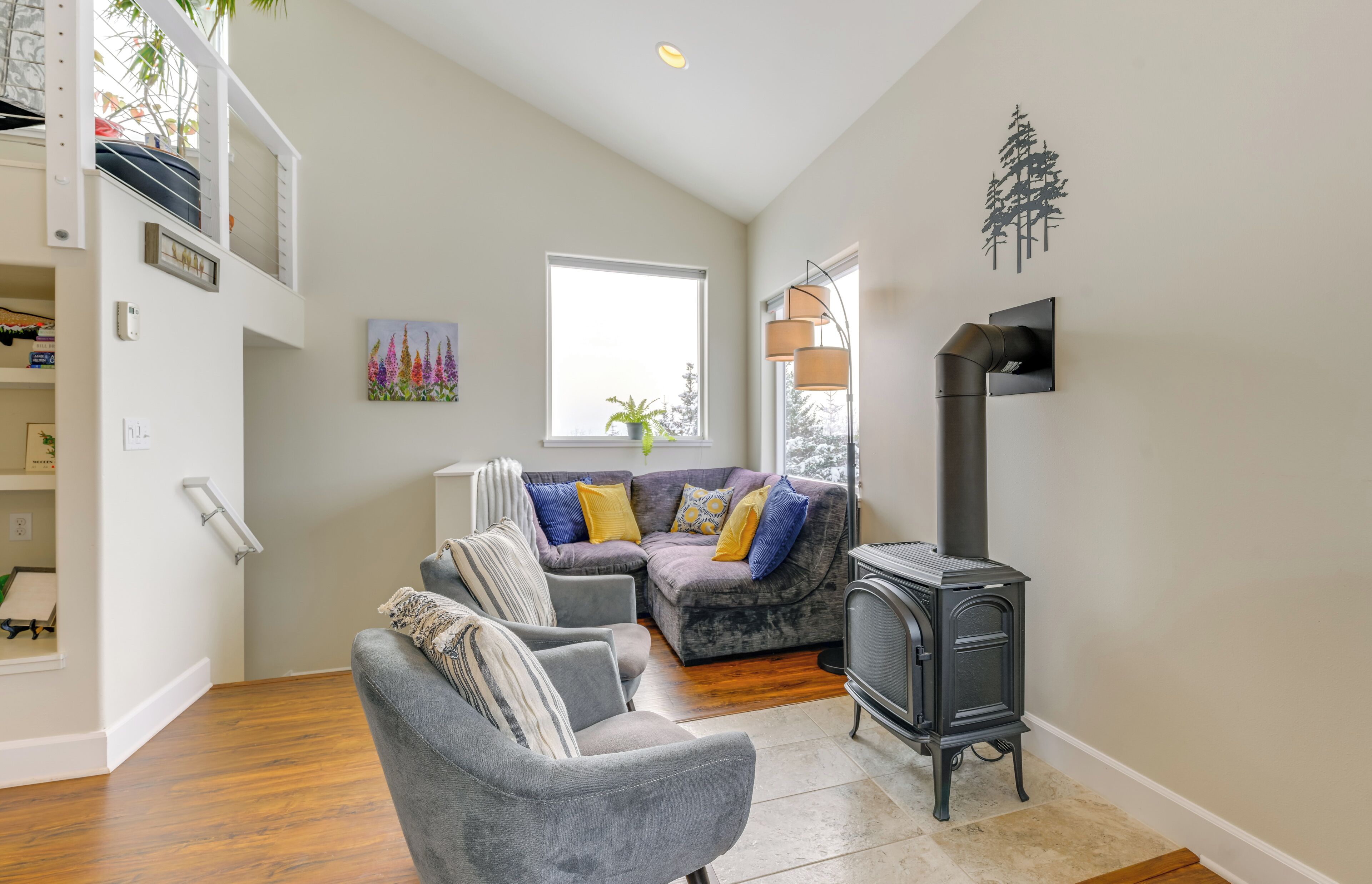 Un salon chaleureux avec un canapé gris, un fauteuil gris et un poêle à bois noir. Une fenêtre offre une vue sur les arbres, et un tableau est accroché au mur.