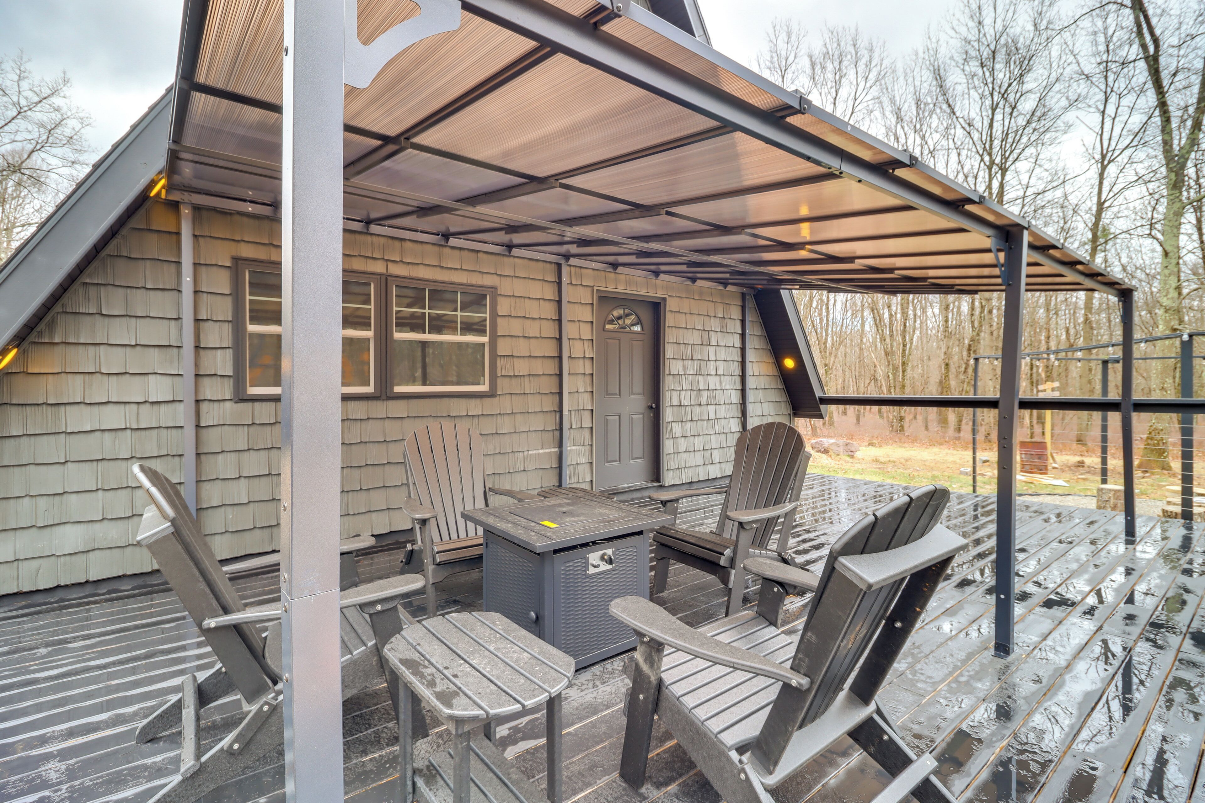 Une terrasse extérieure couverte, équipée d’une table et de chaises, d’un petit radiateur et d’une fenêtre donnant sur une zone boisée.