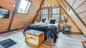 Ein gemütliches Schlafzimmer im Dachgeschoss mit Holzbalkendecke, einem großen Bett mit schwarzer Tagesdecke, einer kleinen Holzbank und einem Wandbild mit Füchsen.