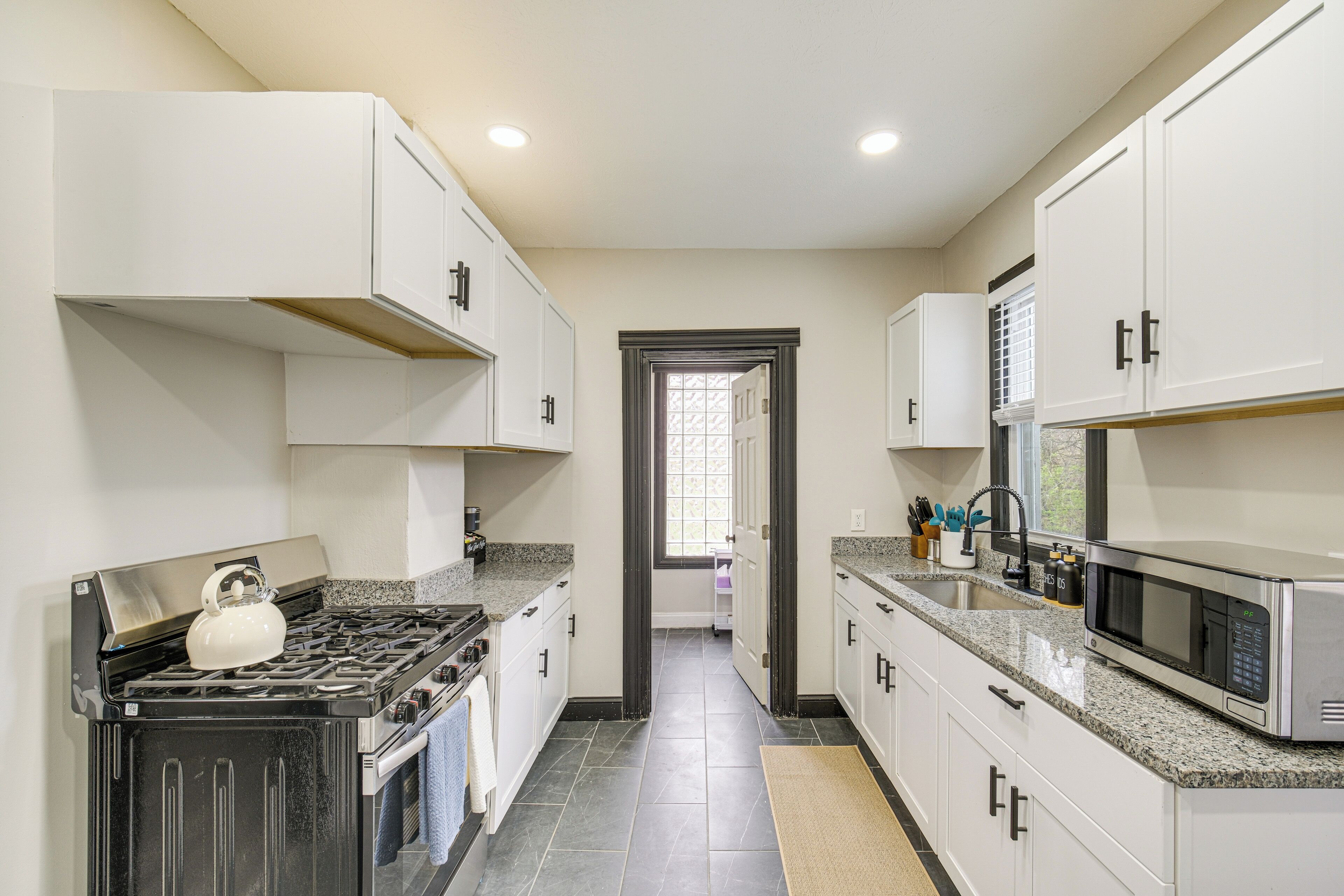 A modern kitchen with white cabinets, a stainless steel stove, a microwave, and a window with blinds.