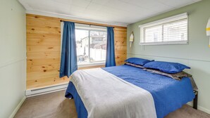 Ein Schlafzimmer mit Holzwand, einem großen Bett mit blauer Bettwäsche, einem Fenster mit Jalousien und Blick ins Freie.