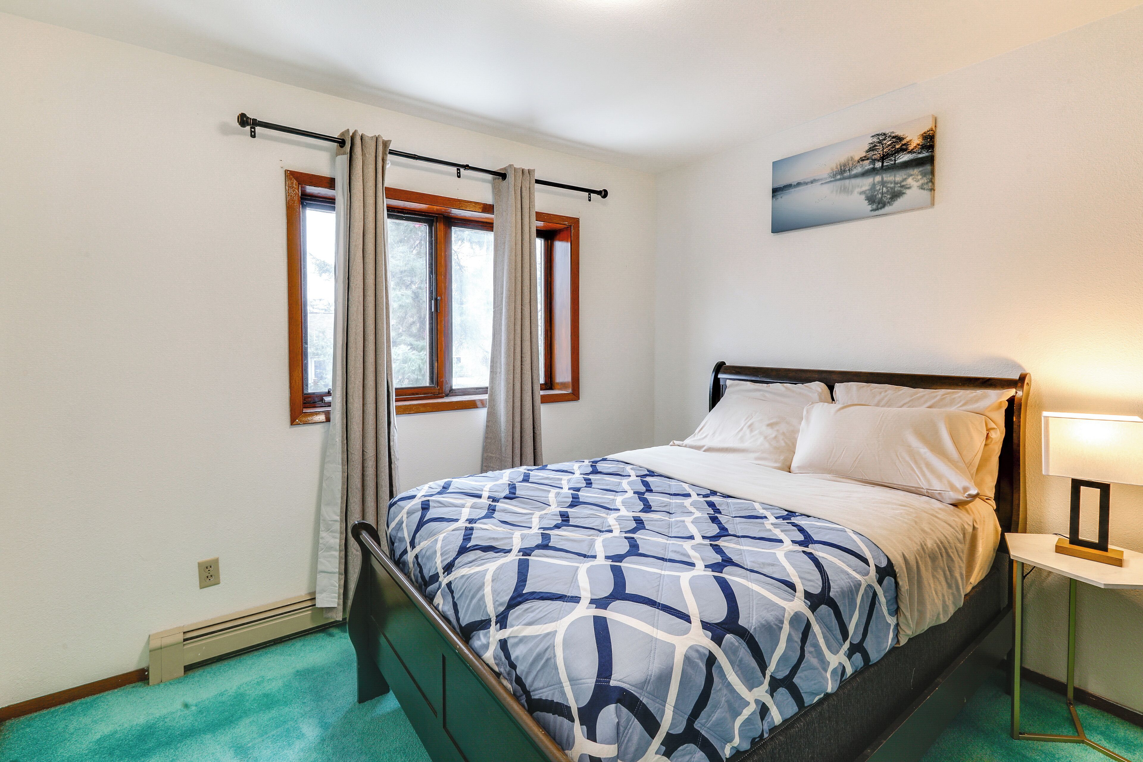 Un dormitorio con cama, ventana con cortinas, mesita de noche con lámpara y un cuadro en la pared.