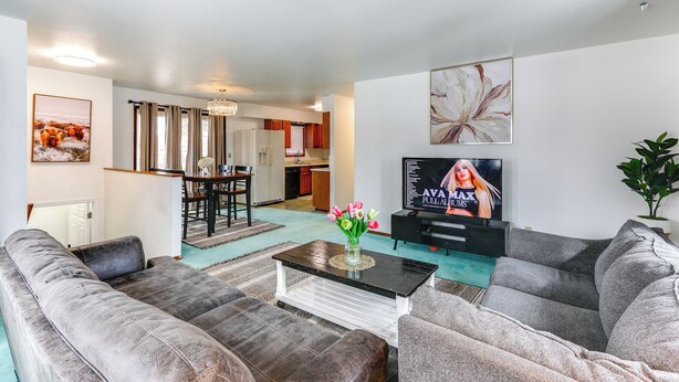 Een moderne woonkamer met een grijze zithoek, een salontafel met een bloevas, een flatscreen televisie, en een eethoek met een kroonluchter.