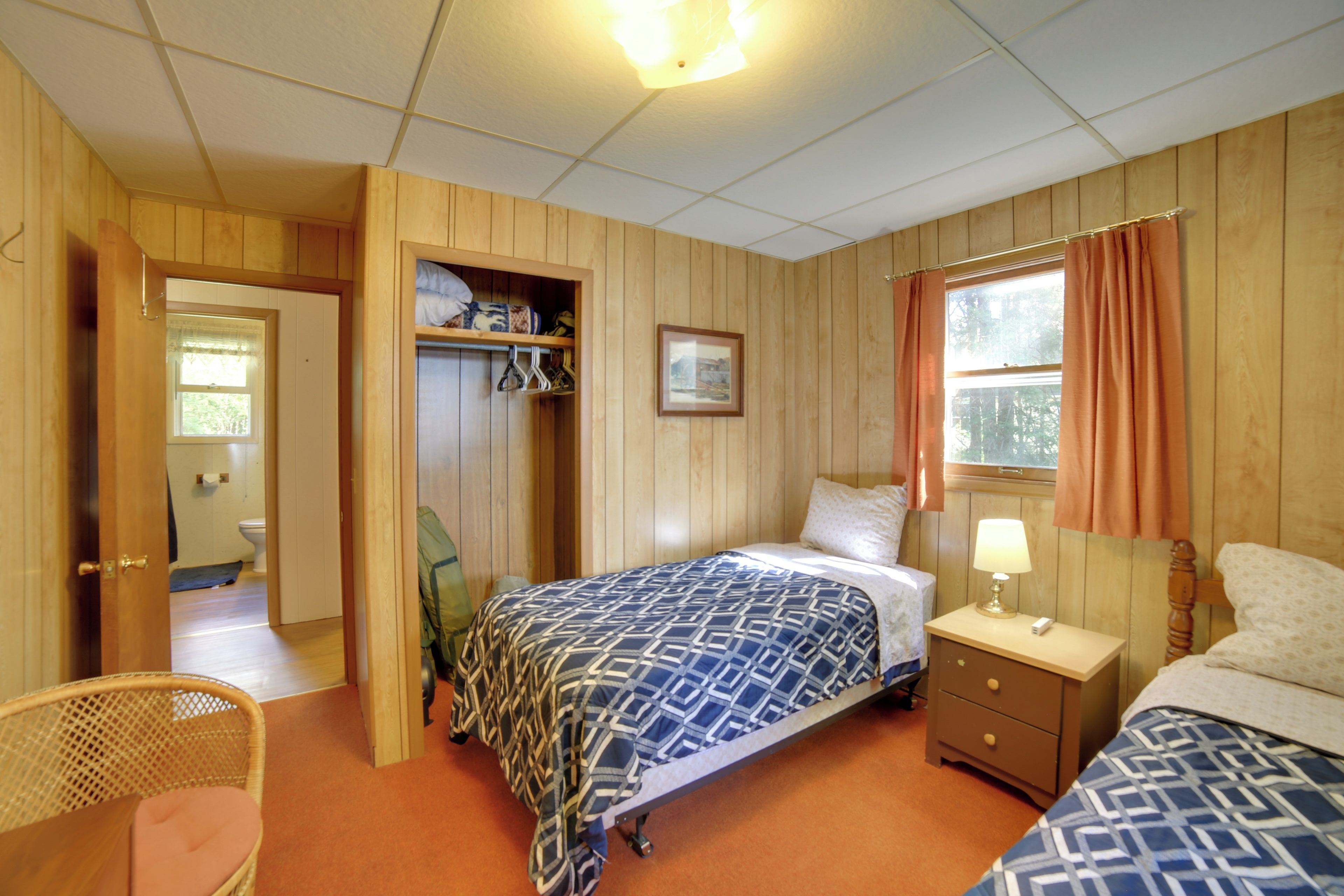 Una habitación con paredes de madera, una cama con ropa de cama estampada, una mesita de noche con lámpara y una ventana con cortinas.