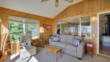 A cozy living room with a sofa, armchair, and coffee table. There is a wooden ladder, a lamp, and a window with blinds.