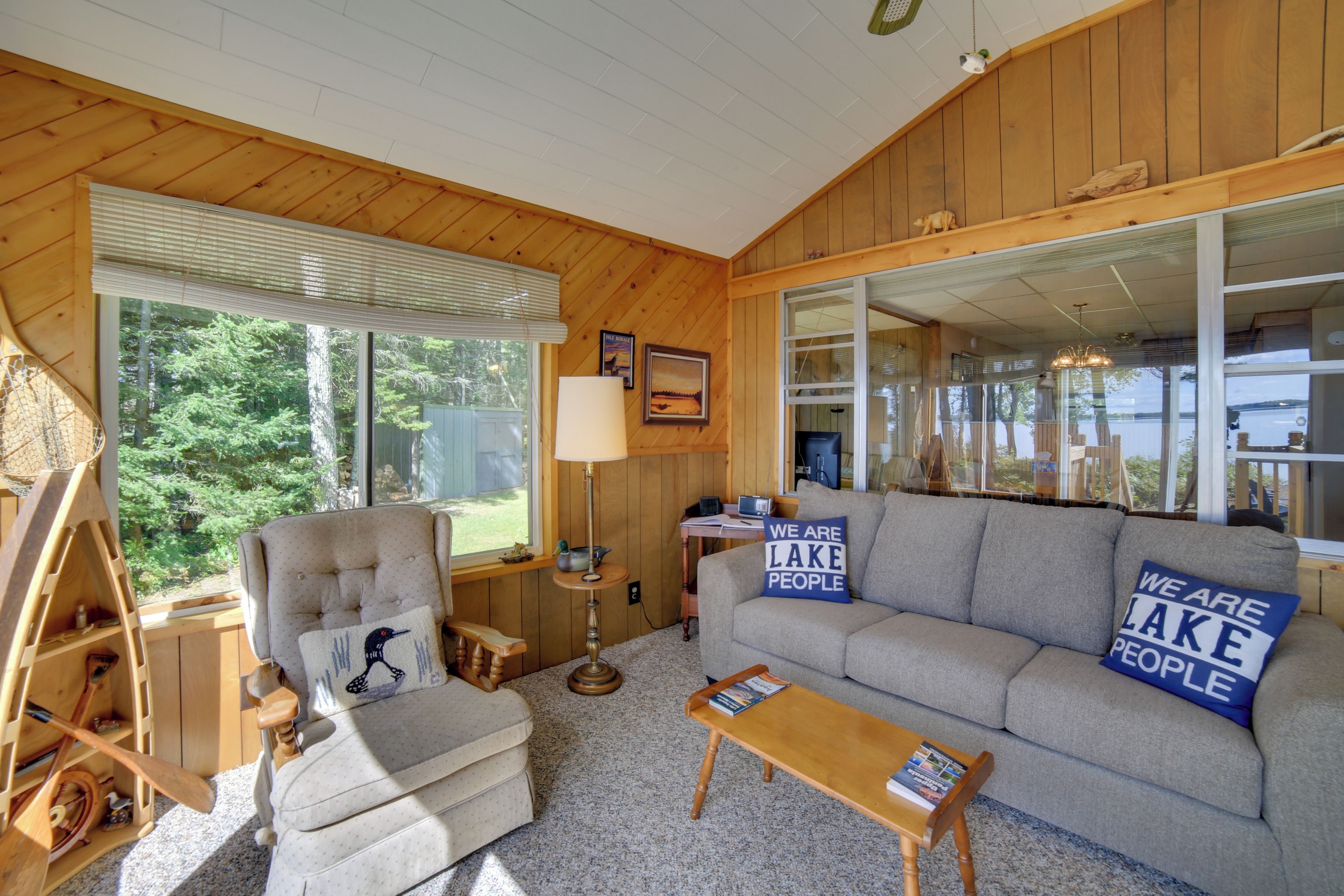 A cozy living room with a wooden interior, a sofa with decorative pillows, a wooden chair, a small table, and a window with blinds.