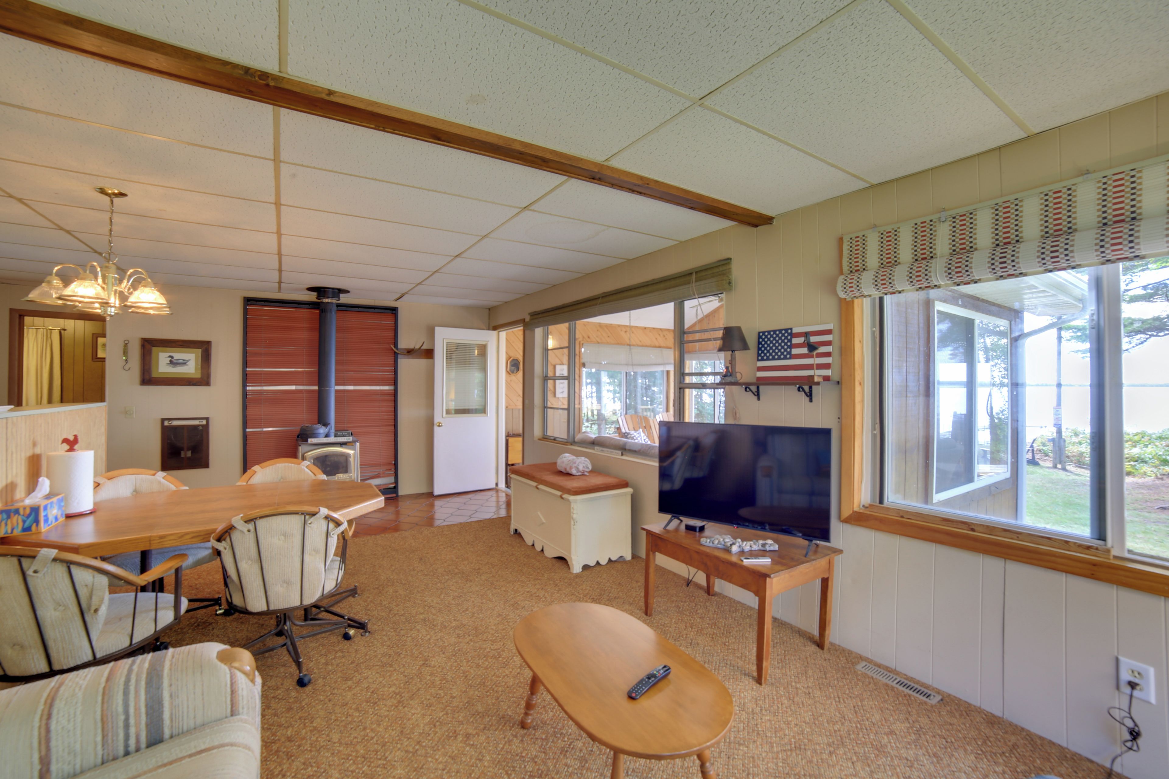 A room with a wooden dining table, chairs, a television, and a window with an American flag.