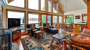 A spacious living room with large windows, a television, comfortable seating, a fireplace, and a kitchen area. - 'sweet Serenity' Schroon Lake Family Retreat! (Schroon Lake)