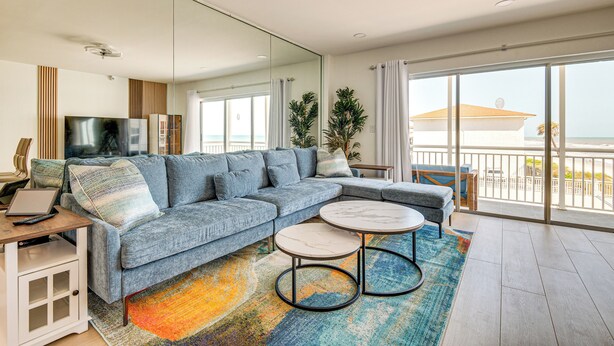 A modern living room with a sectional sofa, a TV, a laptop, and a balcony with a view of the sea. - Steps to Ocean: Satellite Beach Condo w/ Balcony (Satellite Beach)