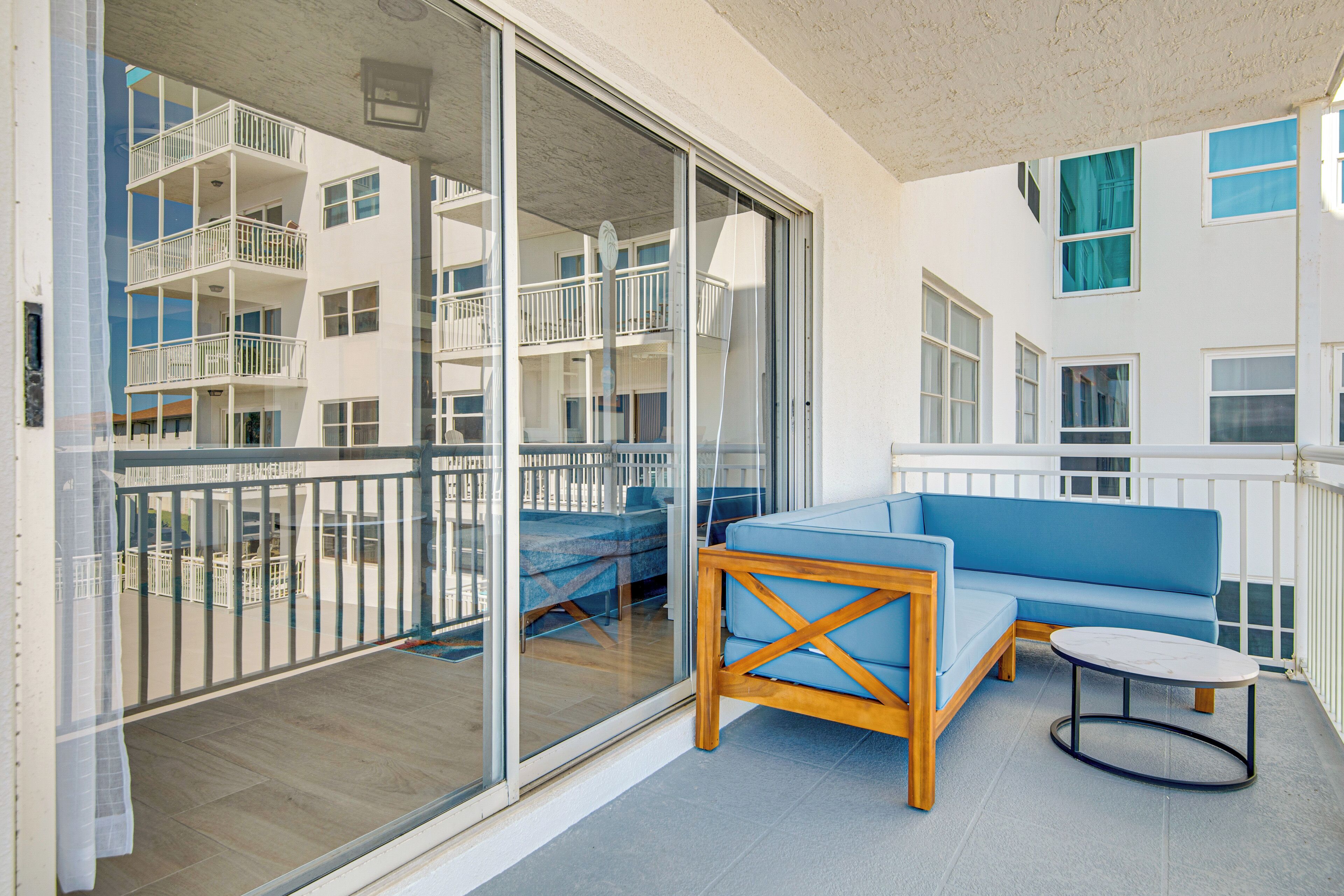 Un balcón con un sofá azul, una mesa de madera y un ventanal de vidrio que da a otro balcón con vistas a edificios.