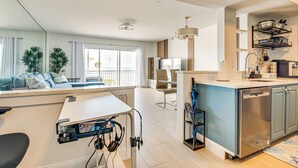 A modern kitchen with a dishwasher, a dining area with a table and chairs, and a living area with a sofa and plants. - Steps to Ocean: Satellite Beach Condo w/ Balcony (Satellite Beach)