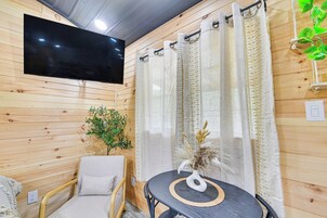 Una habitación acogedora con una pared de madera, un televisor de pantalla plana sobre una mesa redonda con un posavasos tejido, una silla acolchada y un jarrón decorativo con plantas secas.