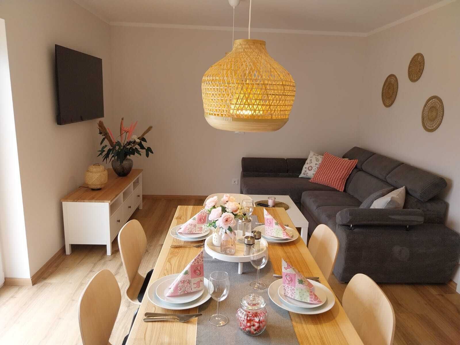 Un coin repas avec une table en bois dressée pour quatre personnes, un canapé gris et un luminaire suspendu en fibres tressées.