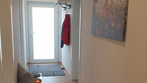 Un couloir avec un plancher en bois, un banc avec une bougie, un porte-manteau avec des vêtements et un tableau accroché au mur.
