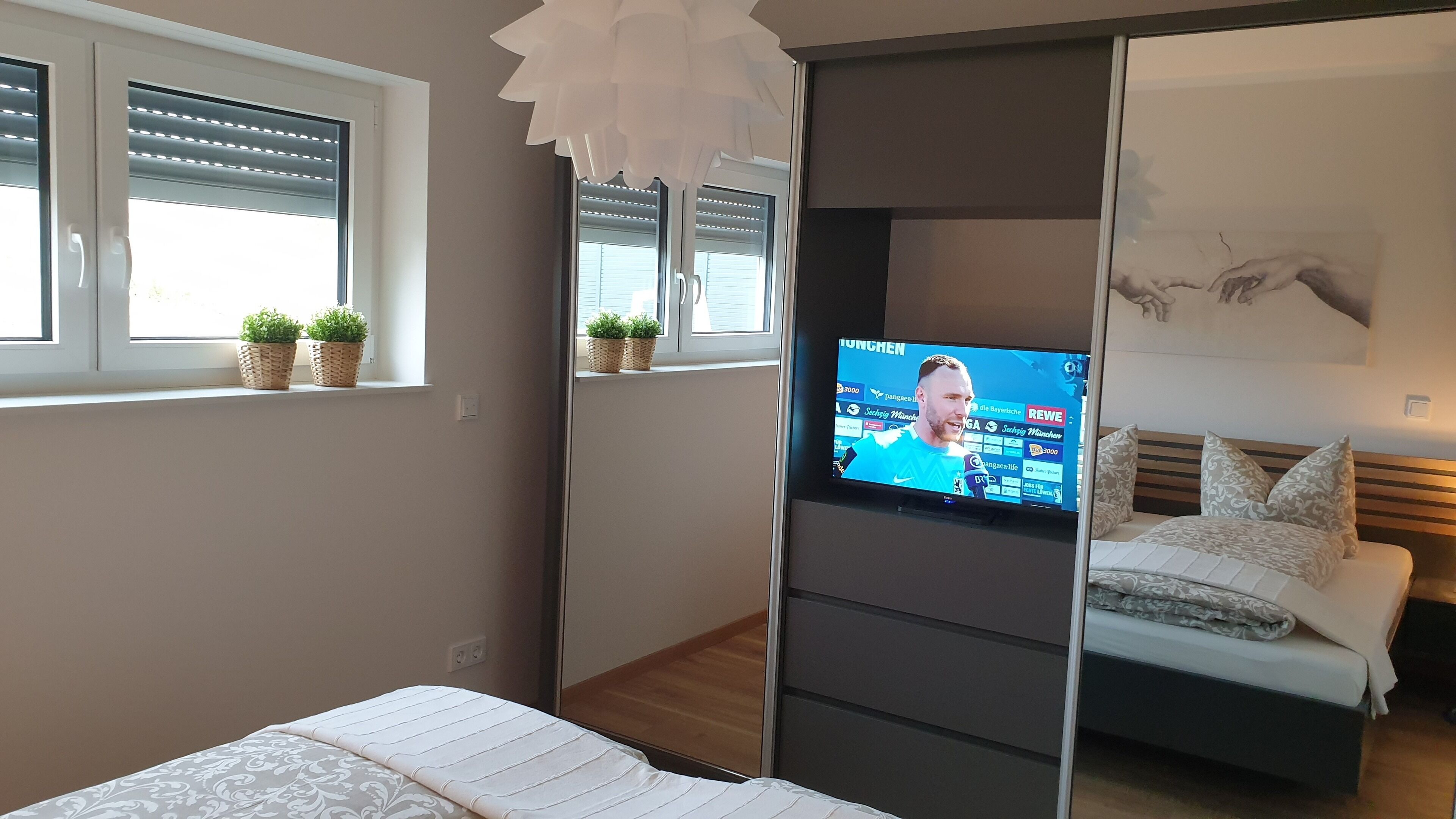 Ein modernes Schlafzimmer mit einem Bett, einem Fernseher auf einem Schrank, einem Fenster mit Jalousien und einer kleinen Pflanze auf der Fensterbank.