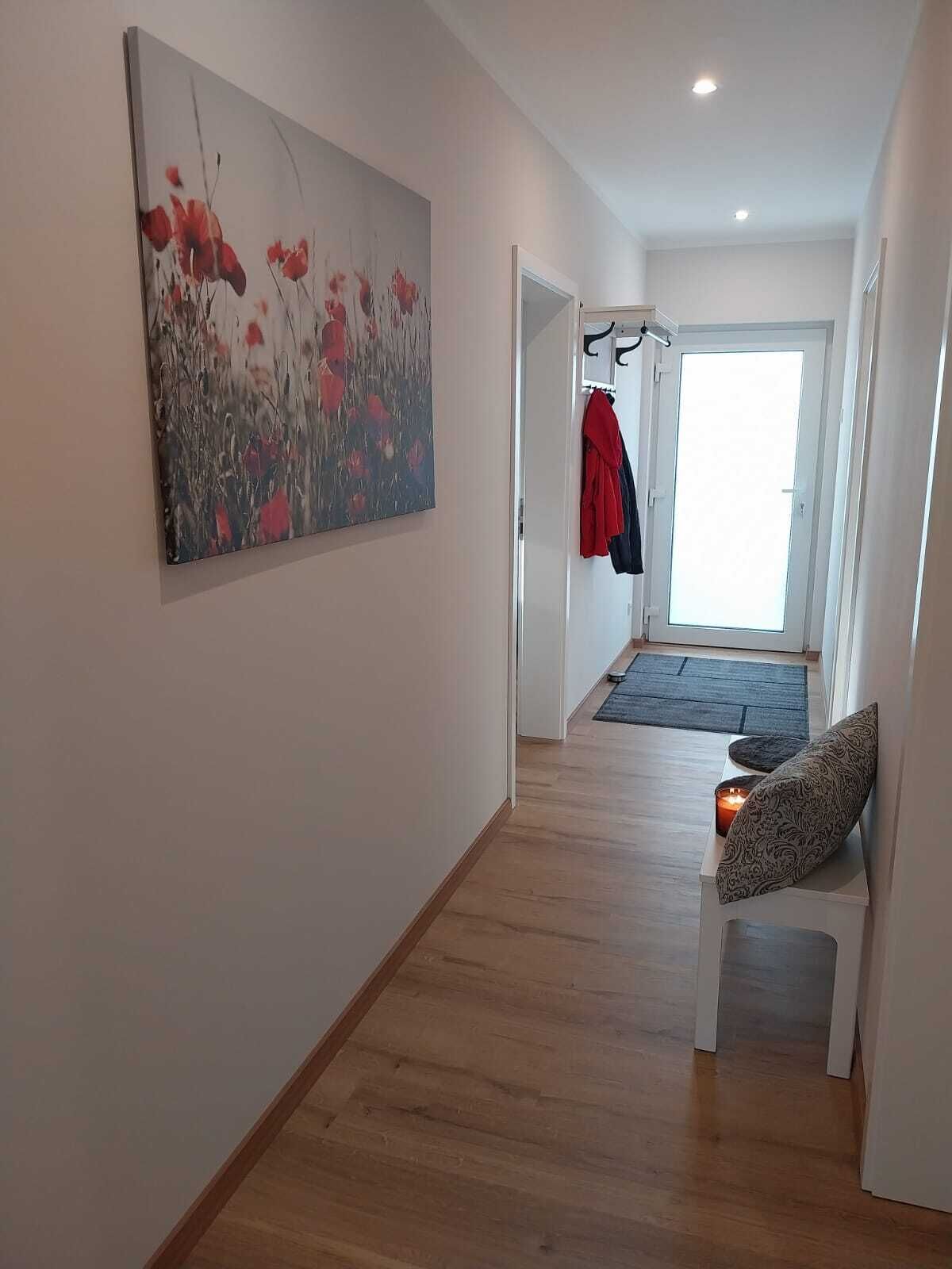Un couloir avec un plancher en bois, un tableau représentant des fleurs rouges au mur, un porte-manteau avec une veste rouge, et un banc avec une bougie.