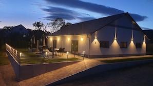 Une maison moderne dotée d’un patio couvert, d’un espace salon extérieur et d’un éclairage extérieur soigné, particulièrement agréable au crépuscule.