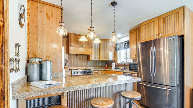 A kitchen with wooden cabinets, a stainless steel refrigerator, a granite countertop, and three bar stools. - 'will's Place' Utah Family Getaway: Hot Tub & Yard (Glendale)