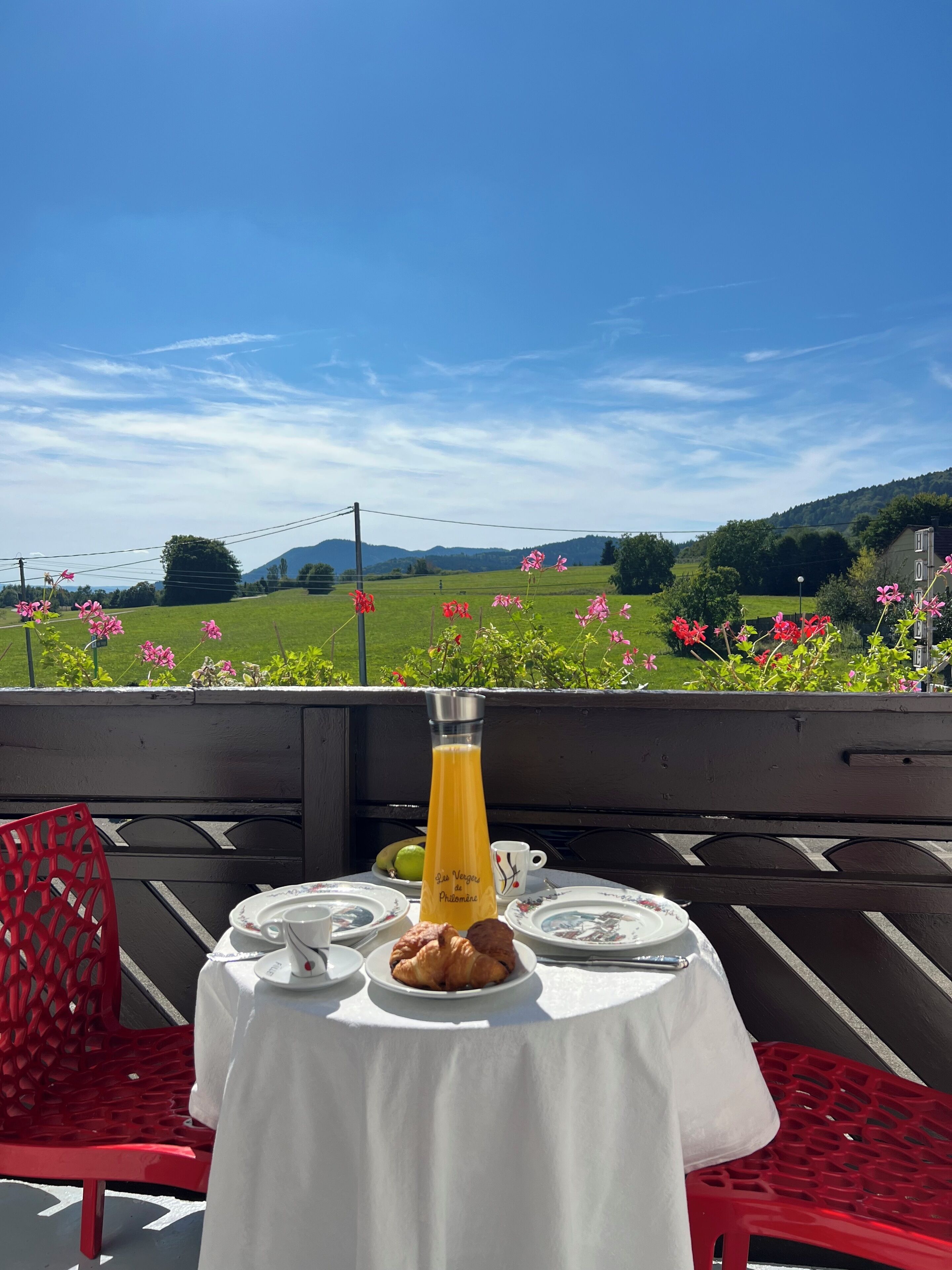 Taula preparada amb un got de suc d'aranja, plats i un croissant a un balcó amb una vista panoràmica de camps verds i flors.