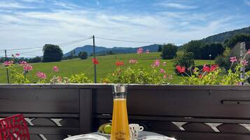 Una mesa dispuesta con jugo de naranja, platos y un croissant en un balcón con vistas panorámicas a campos verdes y flores.