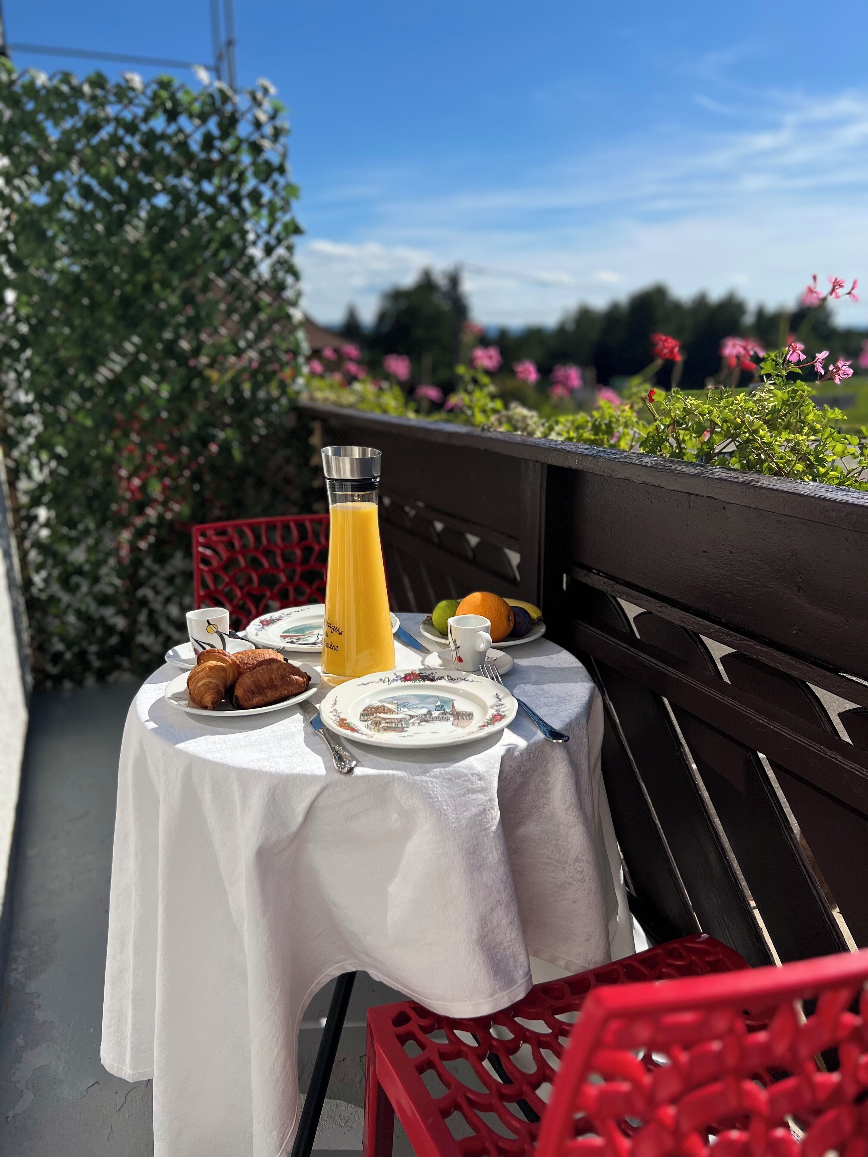 A table set with croissants, a glass of orange juice, and plates with fruits and a cup, on a balcony with a view of flowers and greenery.