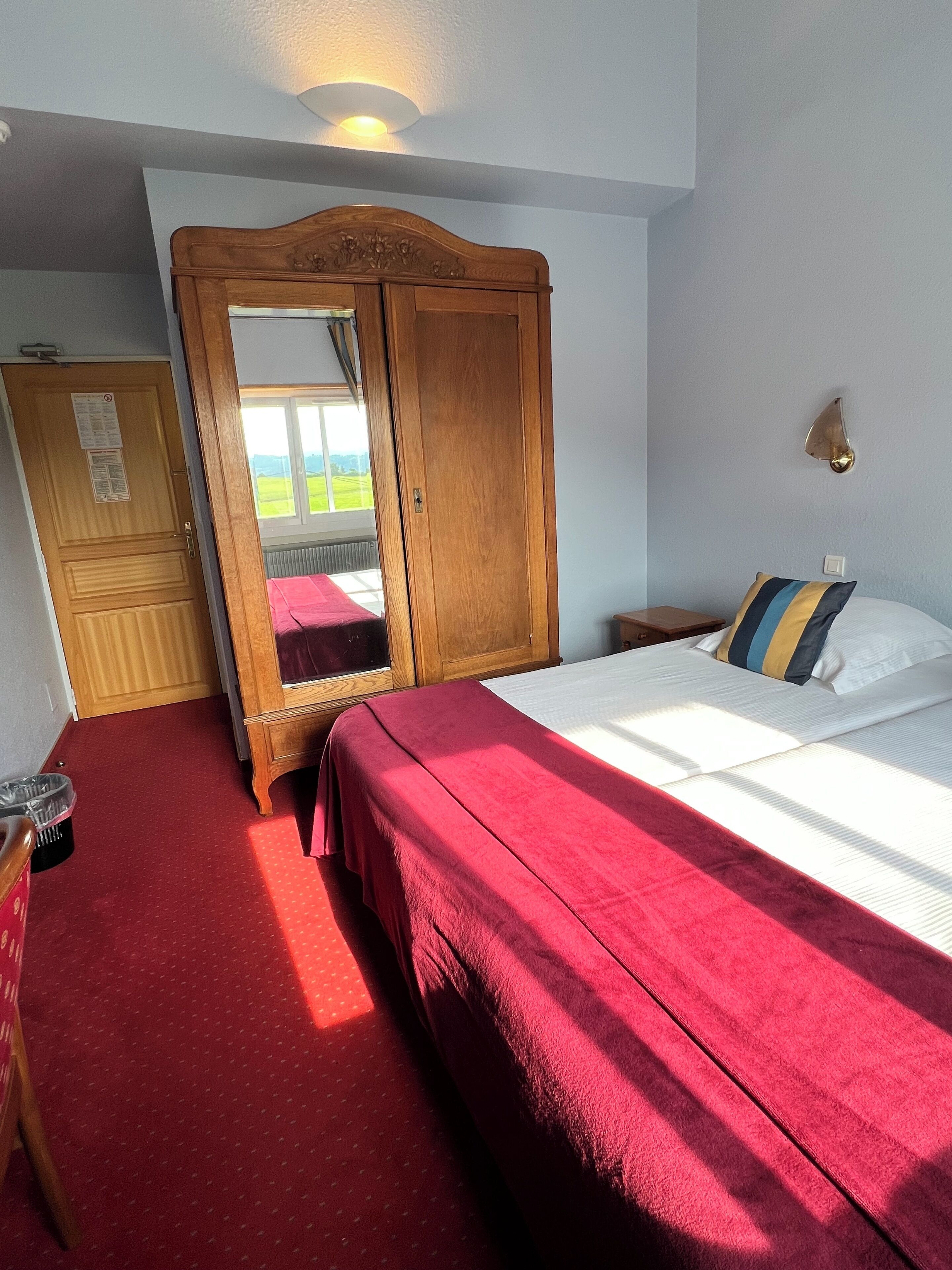 A bedroom with a large bed, a wooden wardrobe, a mirror, and a view of the outdoors.