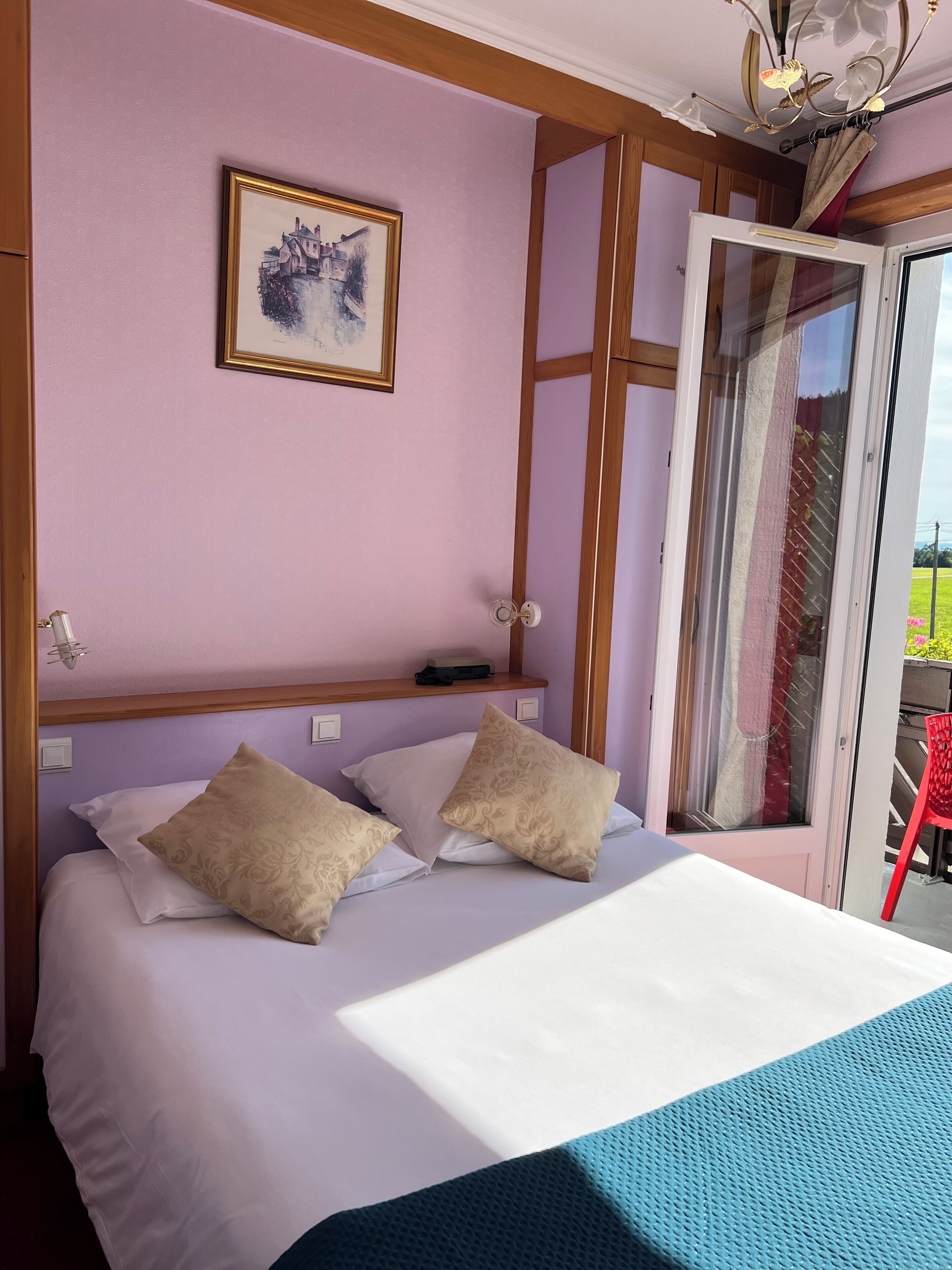 Una camera da letto con un letto, un quadro appeso al muro, una lampada a soffitto e una porta scorrevole che dà sull'esterno.