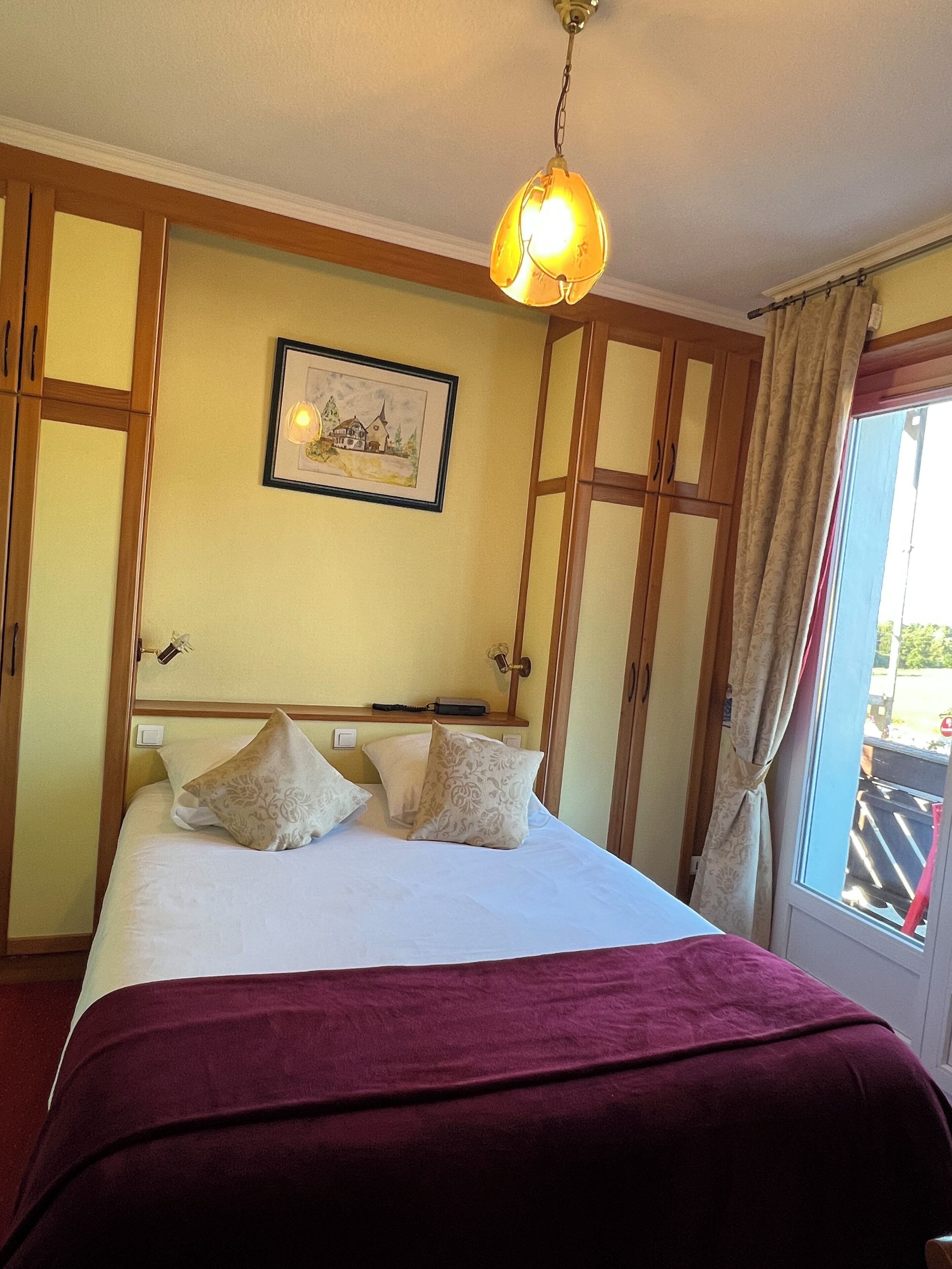 A bedroom with a bed, a hanging light fixture, a framed picture on the wall, and a window with curtains.