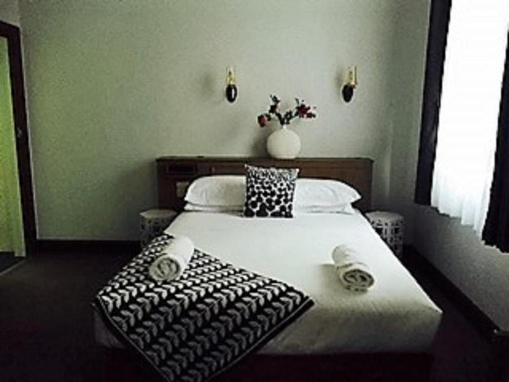 A hotel room with a large bed, two bedside tables, a vase with flowers on the headboard, and a window with curtains.