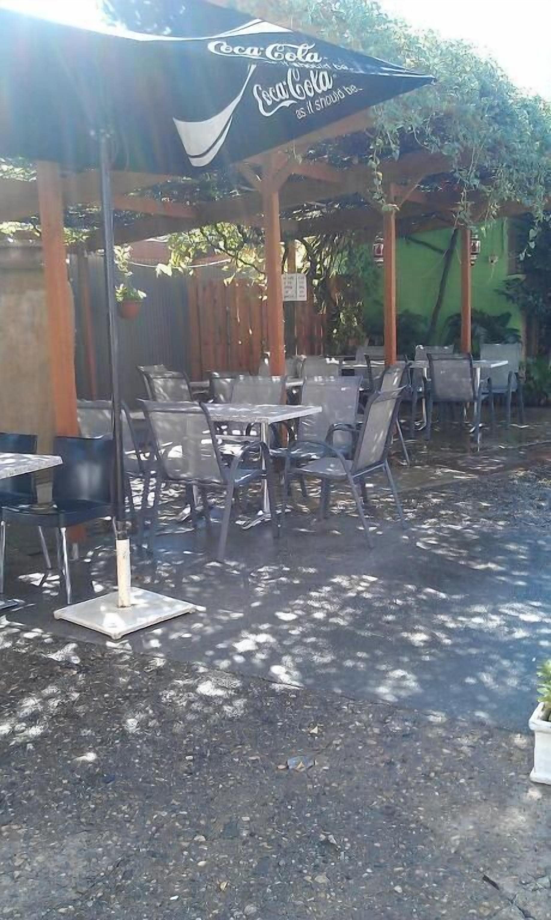 Een terras buiten met tafels en stoelen, een Coca-Cola parasol en een houten pergola.
