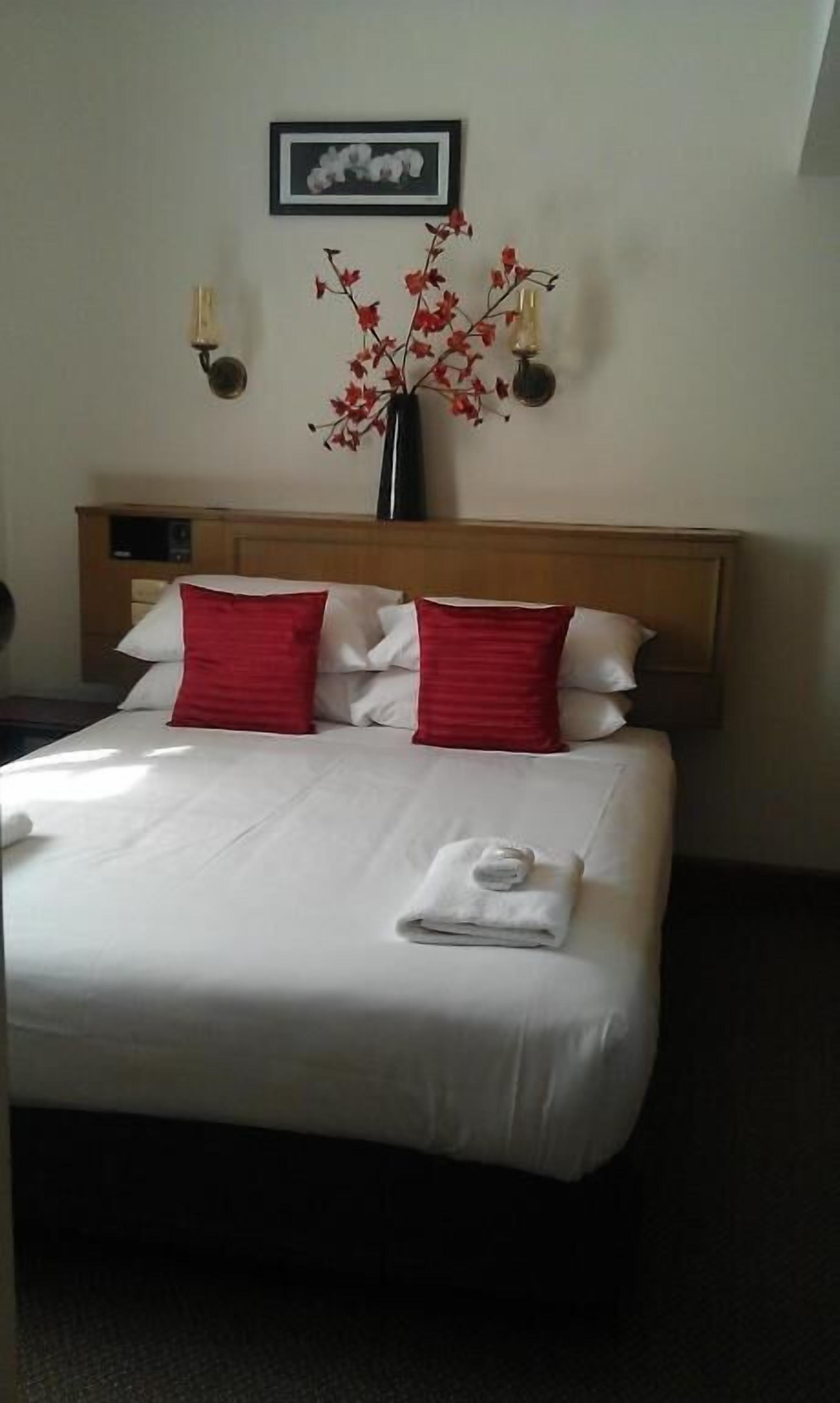 Una habitación de hotel con una cama, almohadas rojas, un florero con flores rojas, un cuadro en la pared y una mesita de noche con una lámpara.