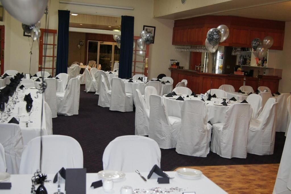 Un salón de banquetes con mesas redondas cubiertas con manteles blancos y sillas, una zona de bar y globos.