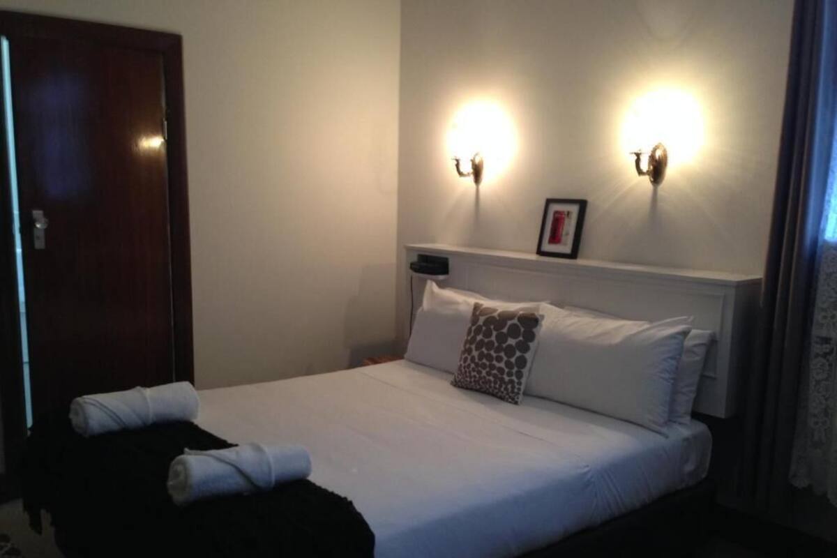 A neatly made bed with white linens and a single patterned pillow, a bedside table with a lamp, and a framed picture on the wall.