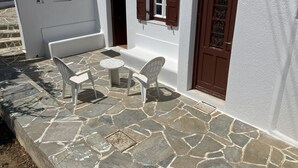 A white building with a stone floor, a small table with chairs, and a brown door. - LINA'S HOUSE (Mykonos)