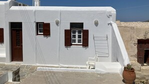 A white building with a stone floor, a door, and two windows with brown shutters. - LINA'S HOUSE (Mykonos)