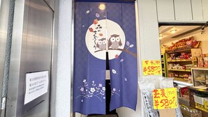 Ein Laden Eingang mit einem blauen Vorhang mit Eulen und Blättern, ein Schild mit japanischem Text und Regale voller Waren.